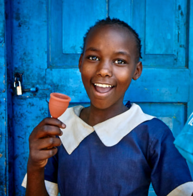 A young girl in school uniform holding a menstrual cup, standing in front of a blue door.